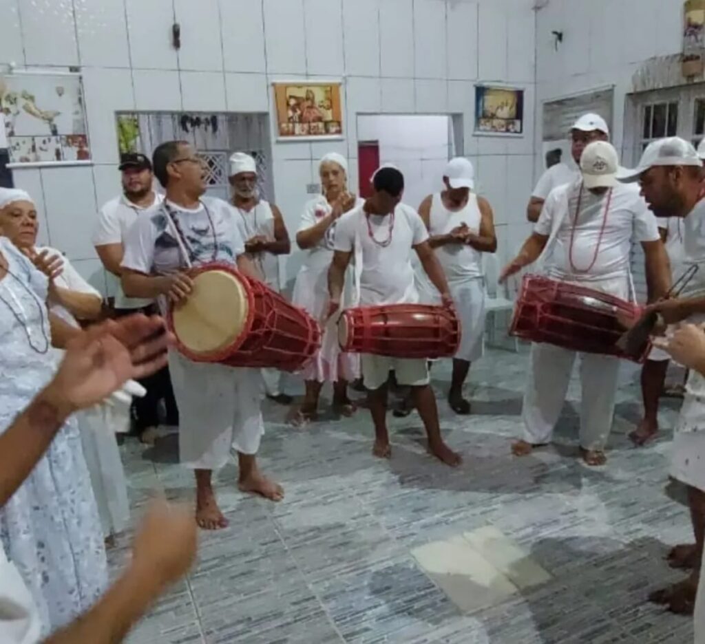 Vivências de Saberes Ancestrais 
Fotografia colorida tirada de um ângulo superior (plongée). Nove pessoas estão reunidas em um círculo dentro de um terreiro com chão de cerâmica cinza. A maioria veste roupas brancas e usa turbantes ou gogós (panos de cabeça) também brancos. Uma das mulheres sentada de costas para a câmera veste uma camiseta com a inscrição "FAMÍLIA BONFIM - Juntos somos mais fortes". Ao fundo, as paredes brancas são decoradas com quadros de entidades religiosas, pequenas máscaras africanas e uma mesa lateral com alimentos e utensílios rituais. O clima é de diálogo e concentração.