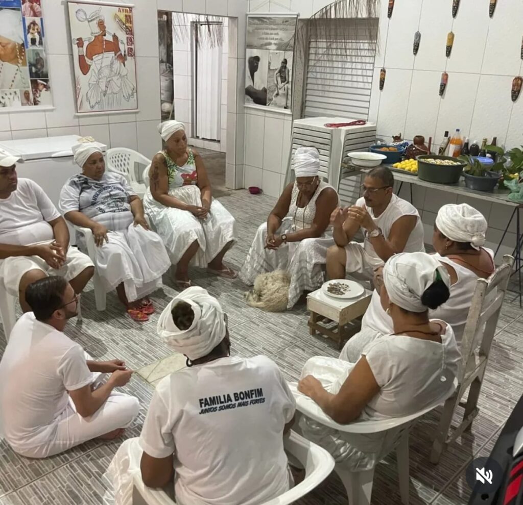 Vivências de Saberes Ancestrais  Fotografia colorida tirada de um ângulo superior (plongée). Nove pessoas estão reunidas em um círculo dentro de um terreiro com chão de cerâmica cinza. A maioria veste roupas brancas e usa turbantes ou gogós (panos de cabeça) também brancos. Uma das mulheres sentada de costas para a câmera veste uma camiseta com a inscrição "FAMÍLIA BONFIM - Juntos somos mais fortes". Ao fundo, as paredes brancas são decoradas com quadros de entidades religiosas, pequenas máscaras africanas e uma mesa lateral com alimentos e utensílios rituais. O clima é de diálogo e concentração.