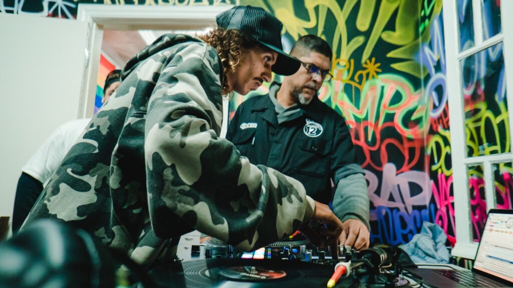 curso extensão hip hip
Fotografia colorida em plano médio. Em primeiro plano, um homem jovem com cabelos cacheados e boné preto, vestindo um casaco de moletom camuflado, opera uma controladora de DJ e um toca-discos. Ele está concentrado, com a mão direita ajustando os controles. Ao fundo, um homem mais velho com óculos observa a ação. O ambiente é decorado com grafites coloridos em tons de verde, rosa e amarelo nas paredes, e há um notebook aberto à direita exibindo um software de música. A iluminação é focada nos equipamentos, criando uma atmosfera de estúdio ou oficina cultural.