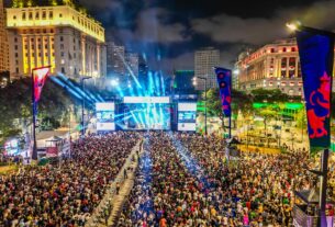 Virada Cultural em uma fotografia aérea noturna de grande escala no Vale do Anhangabaú, São Paulo. A imagem mostra uma multidão compacta de milhares de pessoas ocupando o amplo calçadão central, cercado por edifícios históricos iluminados, como o Prédio da Prefeitura. Ao centro, um palco monumental emite fortes feixes de luz azul e branca em direção ao céu. Dois grandes telões laterais flanqueiam o palco, e estandartes coloridos com artes gráficas verticais estão espalhados pelo local. O horizonte é composto pelas luzes da cidade sob um céu nublado à noite, transmitindo a magnitude do evento cultural.