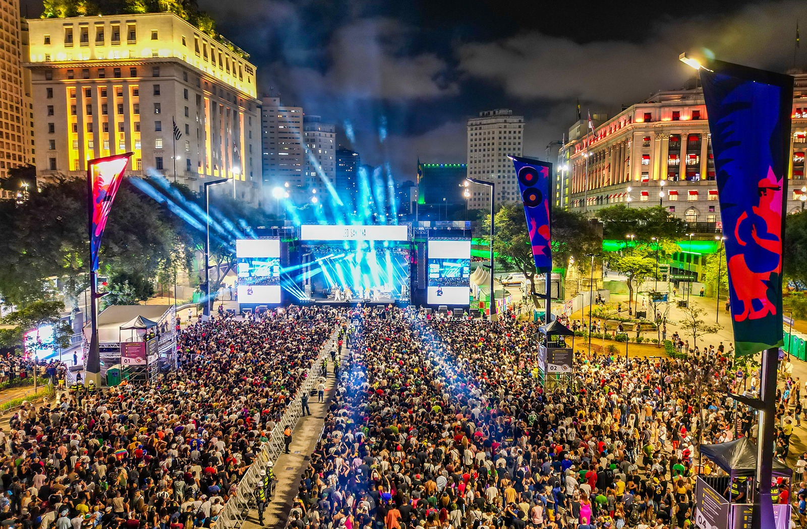 Virada Cultural em uma fotografia aérea noturna de grande escala no Vale do Anhangabaú, São Paulo. A imagem mostra uma multidão compacta de milhares de pessoas ocupando o amplo calçadão central, cercado por edifícios históricos iluminados, como o Prédio da Prefeitura. Ao centro, um palco monumental emite fortes feixes de luz azul e branca em direção ao céu. Dois grandes telões laterais flanqueiam o palco, e estandartes coloridos com artes gráficas verticais estão espalhados pelo local. O horizonte é composto pelas luzes da cidade sob um céu nublado à noite, transmitindo a magnitude do evento cultural.
