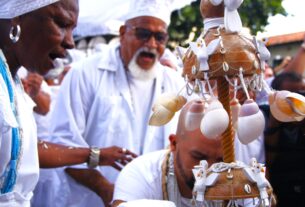 #PraCegoVer: Fotografia colorida em plano médio de uma cerimônia religiosa de matriz africana. Em primeiro plano, à esquerda, uma mulher negra de perfil, com expressão de canto ou clamor, veste branco e colares de contas azuis e brancas. Ao centro, um homem mais velho, com barba branca e óculos escuros, também canta intensamente. À direita, em destaque, um Opaxorô (cajado de Oxalá) feito de madeira, adornado com búzios e pequenas pombas brancas esculpidas. Abaixo, um homem inclina a cabeça em reverência. O grupo veste roupas brancas e o fundo está levemente desfocado, sugerindo uma celebração coletiva ao ar livre.