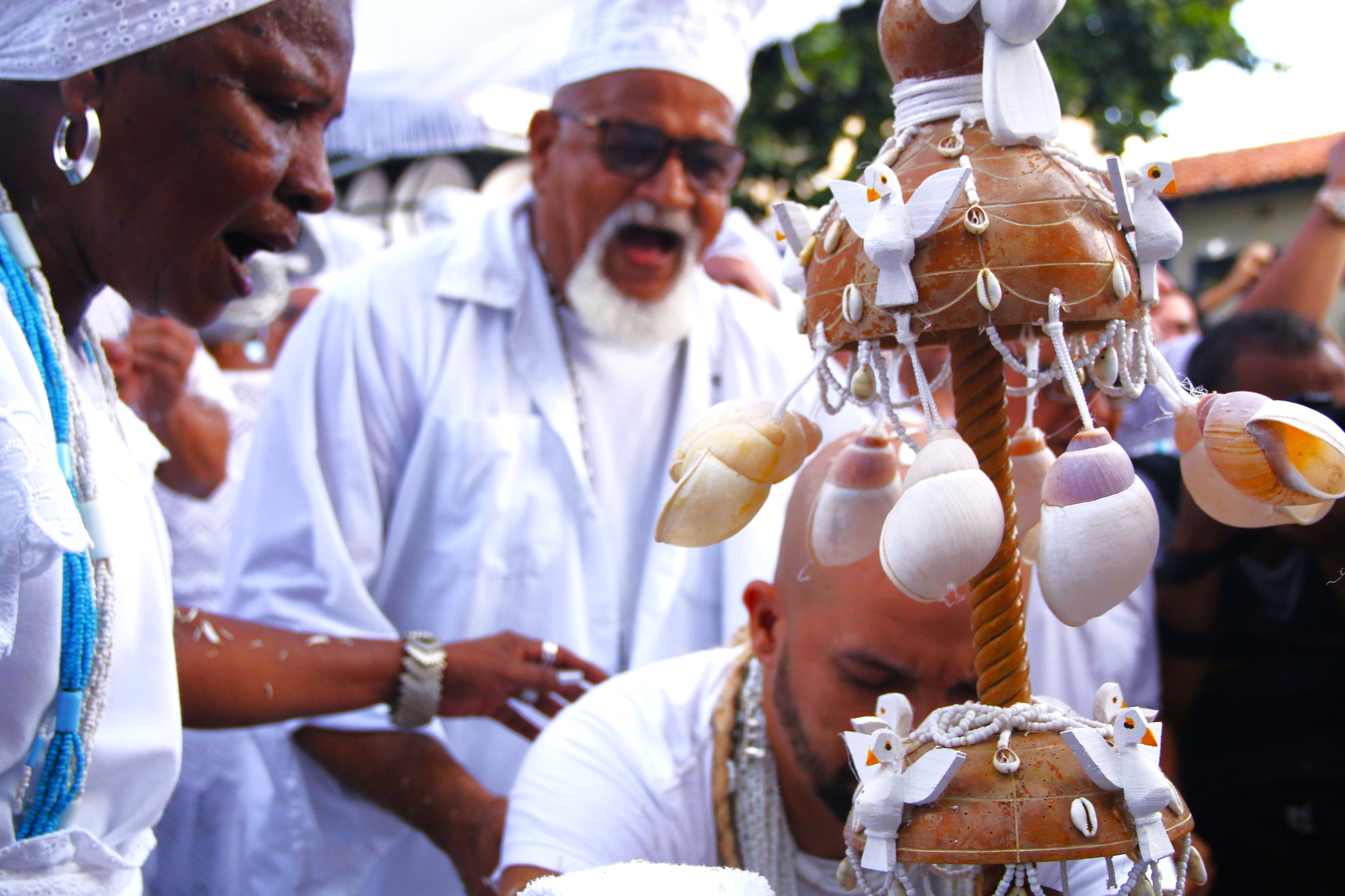 #PraCegoVer: Fotografia colorida em plano médio de uma cerimônia religiosa de matriz africana. Em primeiro plano, à esquerda, uma mulher negra de perfil, com expressão de canto ou clamor, veste branco e colares de contas azuis e brancas. Ao centro, um homem mais velho, com barba branca e óculos escuros, também canta intensamente. À direita, em destaque, um Opaxorô (cajado de Oxalá) feito de madeira, adornado com búzios e pequenas pombas brancas esculpidas. Abaixo, um homem inclina a cabeça em reverência. O grupo veste roupas brancas e o fundo está levemente desfocado, sugerindo uma celebração coletiva ao ar livre.