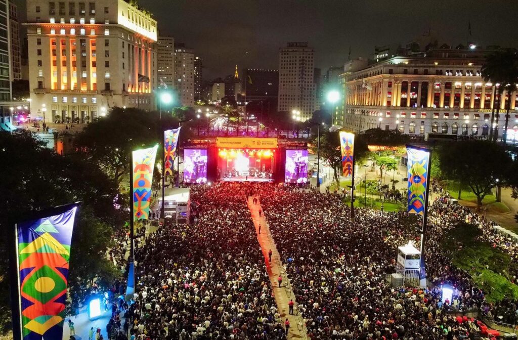 #PraCegoVer: Virada Cultural em uma fotografia aérea noturna de grande escala no Vale do Anhangabaú, São Paulo. A imagem mostra uma multidão compacta de milhares de pessoas ocupando o amplo calçadão central, cercado por edifícios históricos iluminados, como o Prédio da Prefeitura. Ao centro, um palco monumental emite fortes feixes de luz azul e branca em direção ao céu. Dois grandes telões laterais flanqueiam o palco, e estandartes coloridos com artes gráficas verticais estão espalhados pelo local. O horizonte é composto pelas luzes da cidade sob um céu nublado à noite, transmitindo a magnitude do evento cultural.