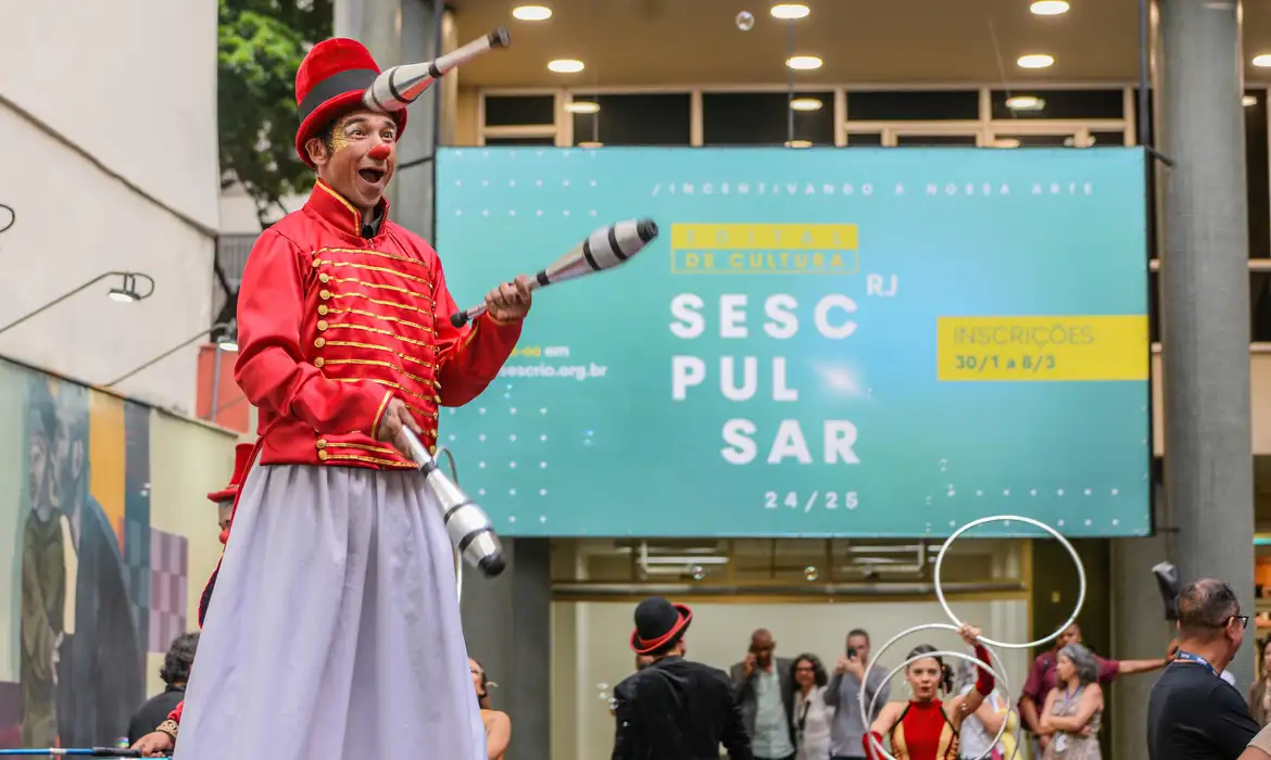 #PraCegoVer:Fotografia colorida em plano médio e aberto. Em primeiro plano, à esquerda, um artista circense sobre pernas de pau sorri com entusiasmo. Ele veste um figurino de mestre de cerimônias vermelho com detalhes dourados, saia longa cinza e um chapéu coco vermelho sobre o qual equilibra um pino de malabares. Na mão direita, ele segura outro pino. Ao fundo, um grande painel azul exibe o texto "Edital de Cultura Sesc Pulsar RJ 24/25", com informações sobre inscrições. No canto inferior direito, outra artista realiza uma performance com bambolês prateados. O ambiente é um pátio externo movimentado com público ao redor. © Adriano Ishibashi/Sesc RJ