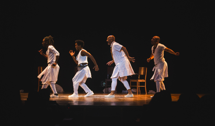 edital sesc pulsar Fotografia colorida em plano médio e horizontal de uma performance teatral. Quatro homens negros estão em cena, alinhados horizontalmente sobre um palco de madeira sob iluminação focalizada. Eles vestem roupas brancas estilizadas, com túnicas e calças que remetem a estéticas ancestrais e contemporâneas. Os artistas estão capturados em movimento, inclinados para a frente como se estivessem em uma caminhada ou dança ritualística. O fundo é um blecaute total (preto), destacando apenas as figuras iluminadas e três cadeiras de madeira vazias posicionadas atrás deles. A cena transmite uma atmosfera de força, ancestralidade e expressão corporal.